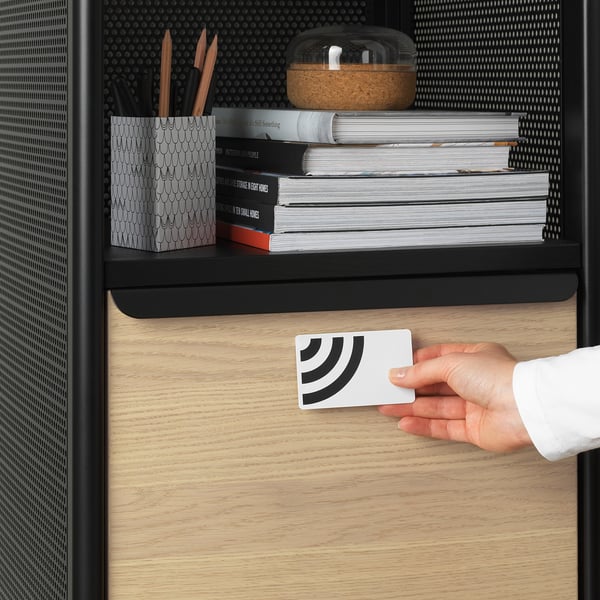 Hand swipes card on wooden desk drawer with black handle, locking mechanism. Above, books and stationery on shelves.