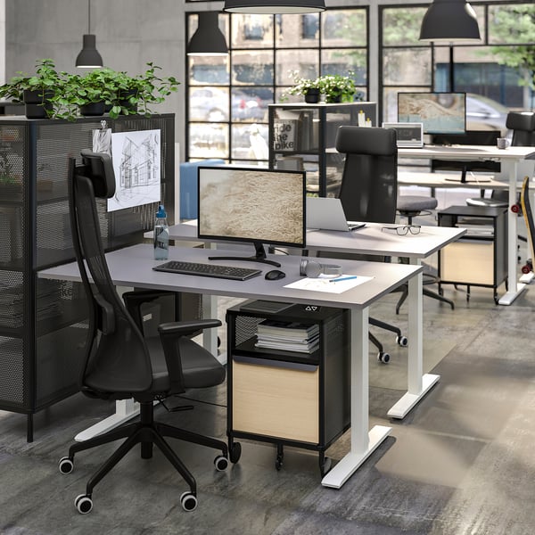 Modern office setup with white BEKANT standing desks, black mesh chairs, and metal storage units. Large windows, plants, and minimalist decor.