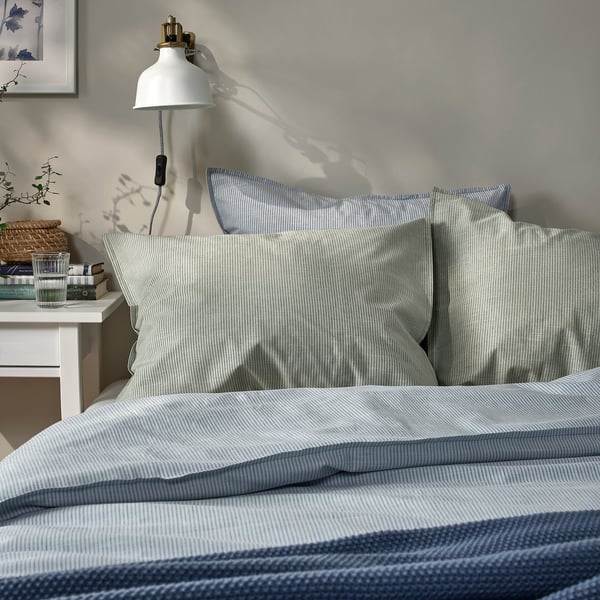 Neatly made bed with light blue striped pillowcases, bedside table, lamp, books, glass; cosy, compact bedroom.