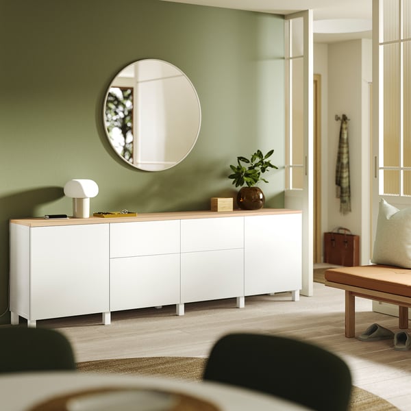 Modern living room with BESTÅ white sideboard, round mirror, green wall, wooden bench, and potted plant.