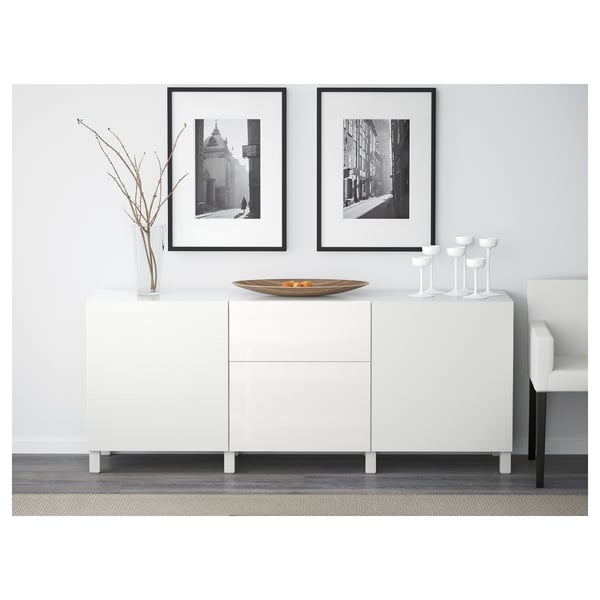 White sideboard with drawers and shelves. Framed black-and-white art above. Bowl, glasses, and decorative vase on top.