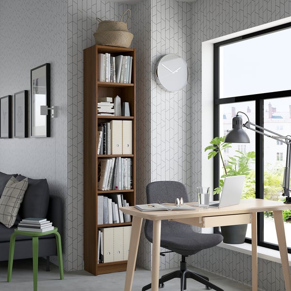 A well-organized home office with a wooden bookcase, office chair at a desk by a window with a laptop and plant, and a cosy sofa with pillows.