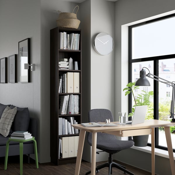 Modern home office: tall BILLY bookcase with books, grey chair, small green stool, cosy couch, desk with laptop, large window.