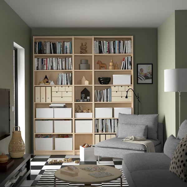 Large wooden BILLY bookcase fills corner, packed with books, decor items. Grey sofa and patterned rug nearby.