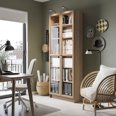 Light wooden BILLY bookcase with glass doors, near a white chair & small table, decorated with plates and vases.
