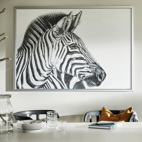 Large framed black and white zebra portrait, BJÖRKSTA, hangs above a white table with chairs and items.