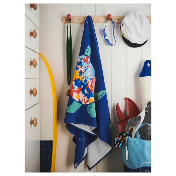 A colourful turtle-patterned blue bath towel hangs on a wooden hook next to a white wall and drawers with wooden knobs.