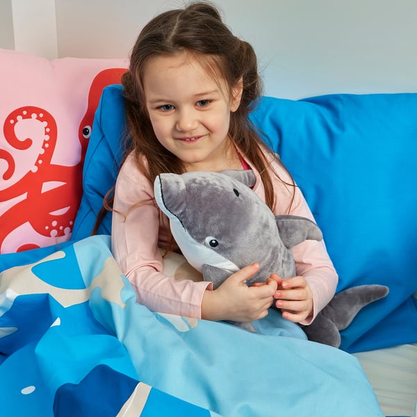 A young girl hugs a grey dolphin cuddly toy on a blue bed. Shes in pink pyjamas under a blue blanket. A red octopus pillow is behind the person.