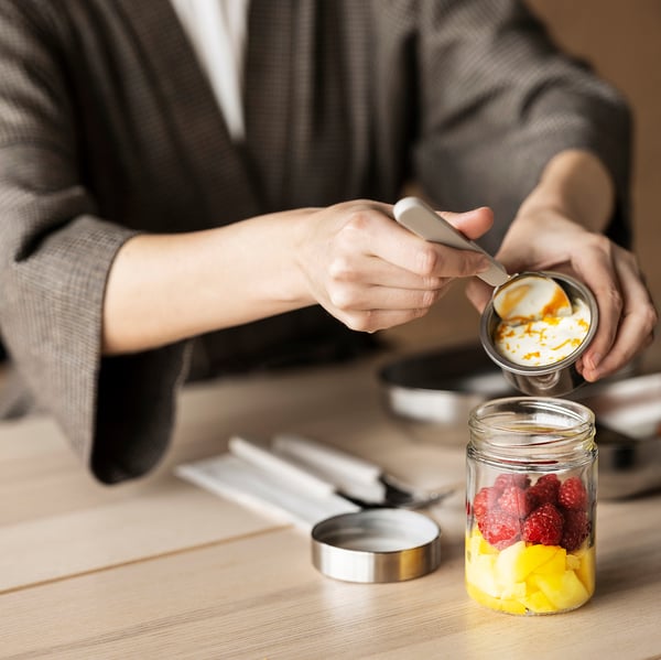 A person makes a fruit and yoghurt parfait in a DAGKLAR jar with a separator on a wooden table among kitchen tools.