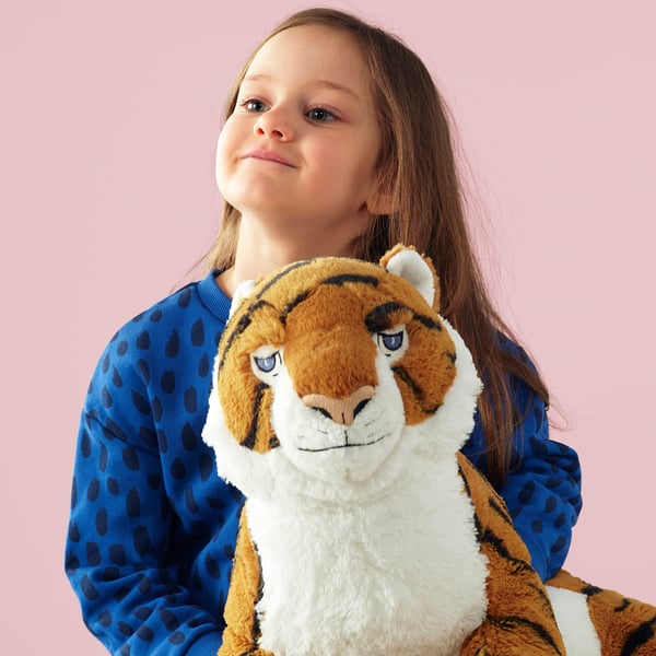 A girl in a blue dotted outfit holds a large plush tiger toy, smiling upward. The tiger has stripes and a white face.