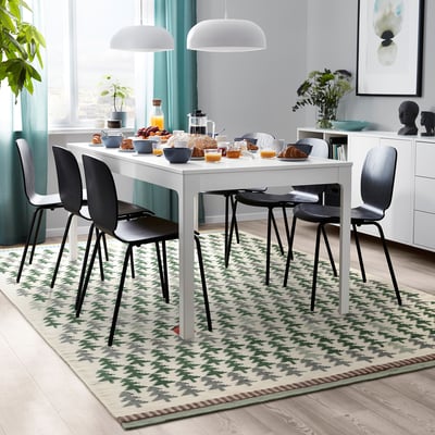 Cosy dining area with rectangular white table, four black chairs, patterned green rug. Breakfast items on table under white pendant lamps.