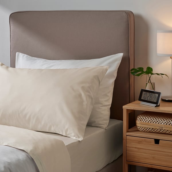 Bedroom scene with light-colored DVALA bedding. Pillowcase and sheets are 100% cotton. Wooden headboard and bedside table, clock and plant nearby.