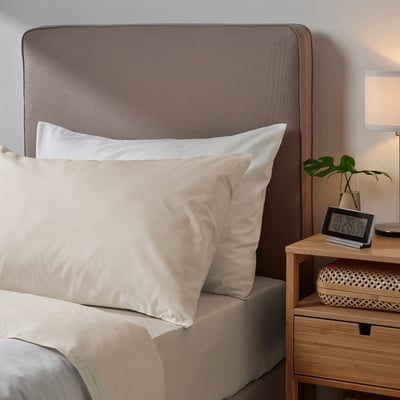 Bedroom scene with light-colored DVALA bedding. Pillowcase and sheets are 100% cotton. Wooden headboard and bedside table, clock and plant nearby.