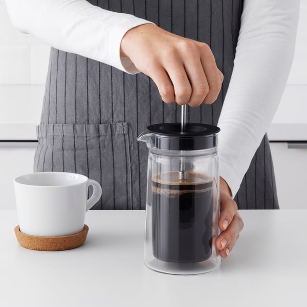 Person presses down the plunger on a glass cafetière, extracting coffee grounds into a dark brew. A white mug waits nearby on a cork coaster.