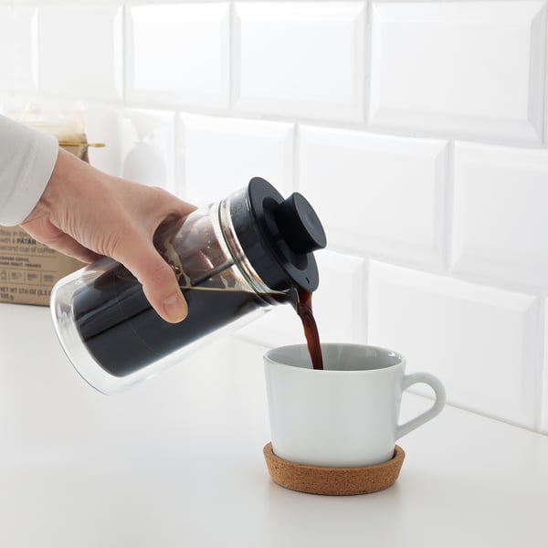 A hand pours freshly made coffee from a glass press into a white mug, sitting on a cork coaster.