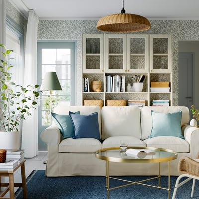 Bright living room with light-colored EKTORP sofa, blue pillows, glass coffee table, bookshelf, and large window.