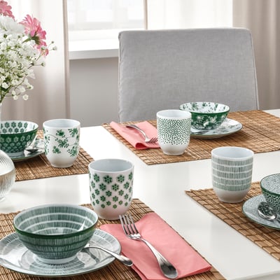 Table set with green-patterned ENTUSIASM mugs and bowls, pink napkins, utensils, and flower vase near window.