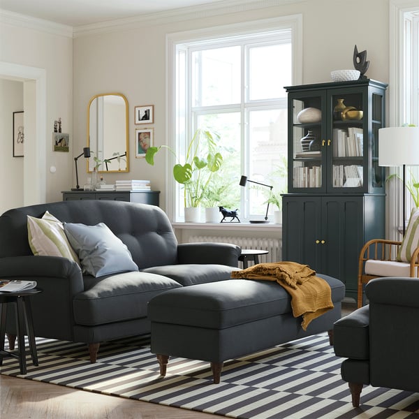 Modern living room with large grey sofa, striped rug, gold mirror, dark green bookcase, plants, and cosy decor.