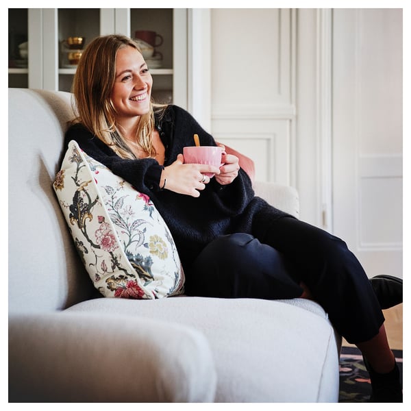 Person smiles, sitting on white ESSEBODA sofa, holding pink mug. Floral pillow, black attire.