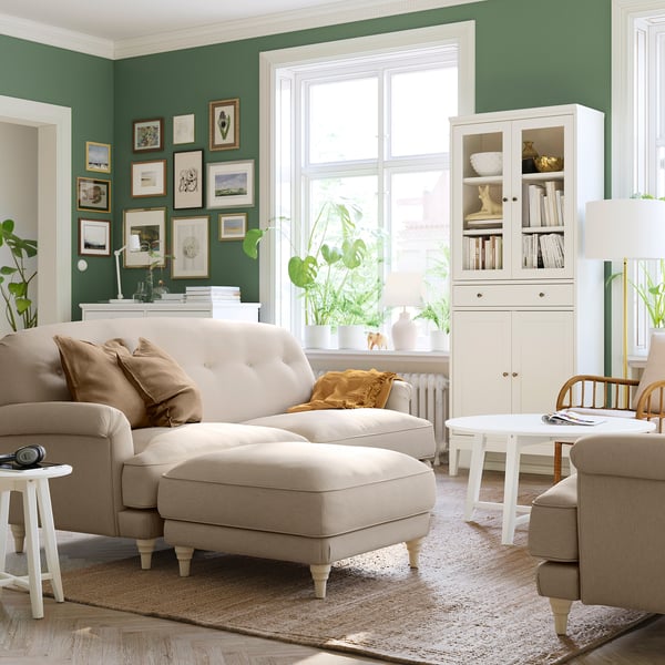 Modern living room with green walls, beige sofa, ottoman, and white cabinet. Natural light from large window, plants, and cosy decor.