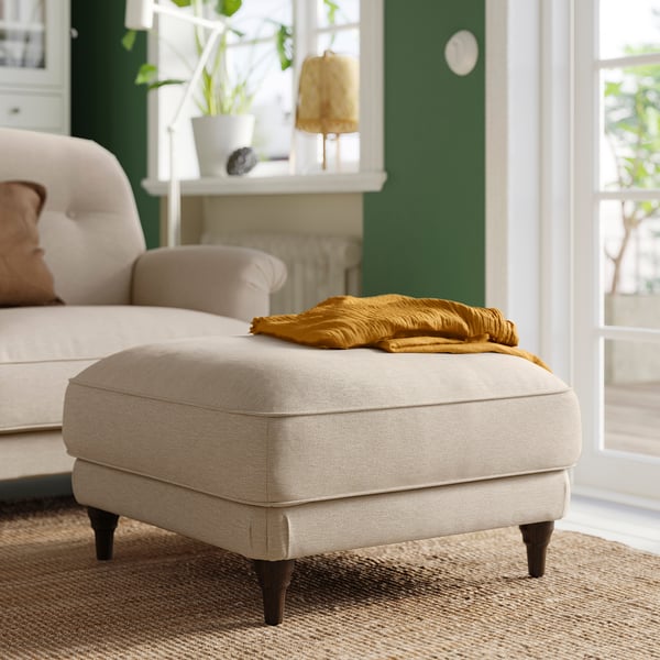 Cosy living room with ESSEBODA beige footstool, soft blanket, green wall, white shelf with plants, and natural rug.