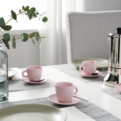 A dining table set with pink cups and saucers on placemats, beside a silver coffee maker, with chairs around.