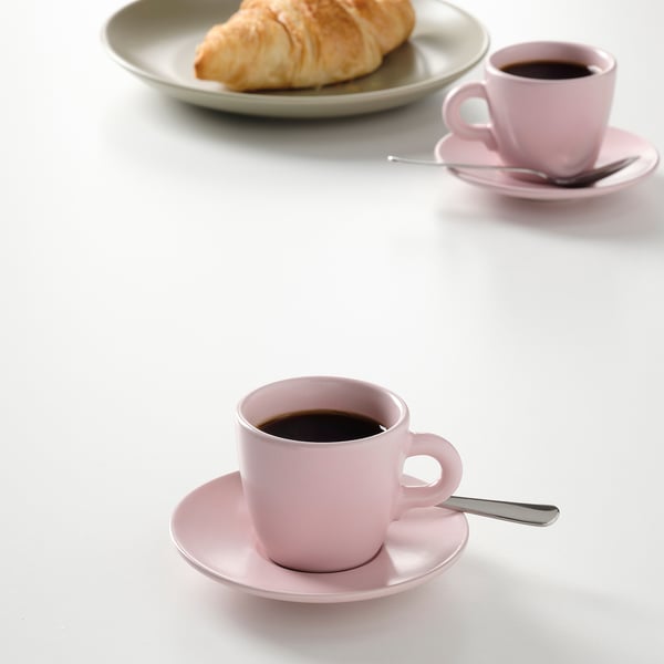 Two pink coffee cups with saucers, one with a spoon and a pastry on a plate in the background.