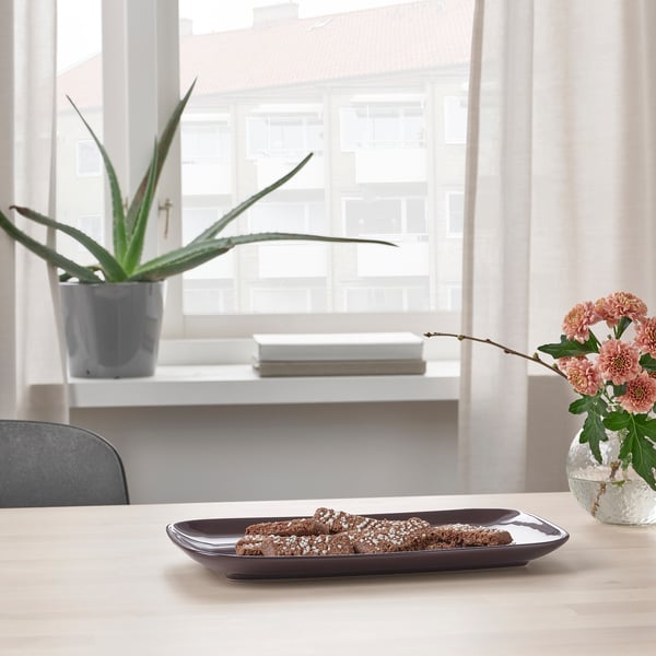 A wooden dining table with a rectangular plate holding snacks, situated near a windowsill adorned with a potted plant and a vase of pink flowers.