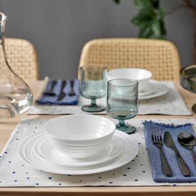 FAVORISERA dinner set on blue cloth, with stackable plates, bowl, narrow-stem glasses & silverware. Wicker chairs in background.