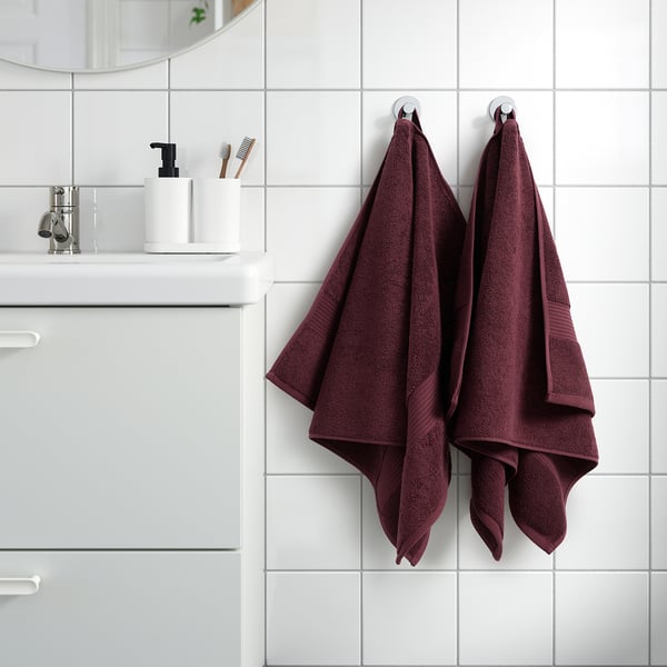 Maroon FREDRIKSJÖN towels hang on bathroom hooks, surrounded by minimal decor with white tiles and vanity, showcasing soft cotton texture.