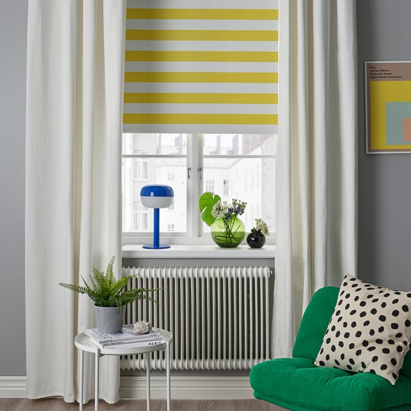 Modern living room with yellow-striped blind, blue lamp, vases, green chair & round table plant.