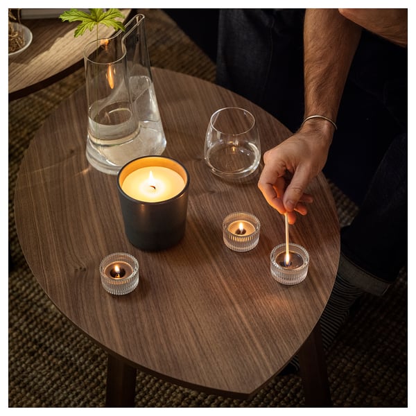 Person lights candles on wooden table: large scented and small tea lights, empty glasses, vase with plant. Cosy lighting.