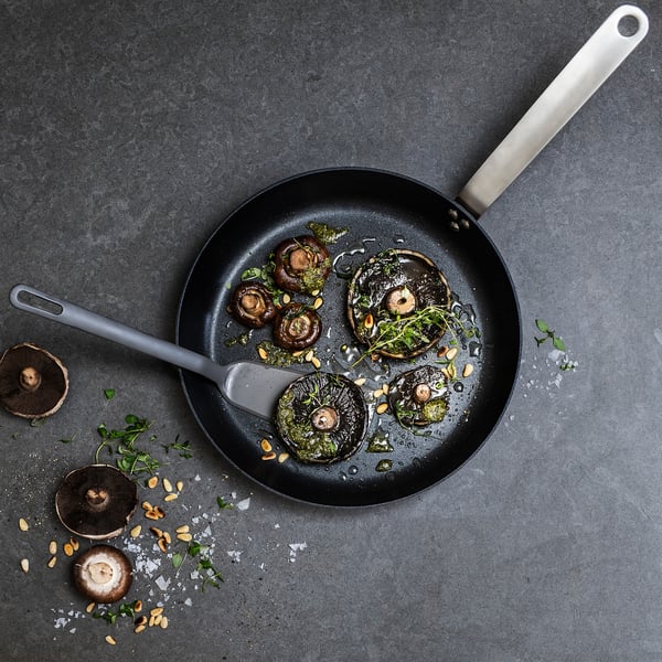 A black pan with a grey handle, containing cooked mushroom slices and herbs. Surrounded by scattered mushrooms and almonds on a dark surface.