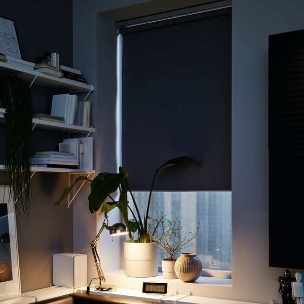 Modern black FYRTUR roller blind on window, blocking light, part of home office with plants, books, and lamp.
