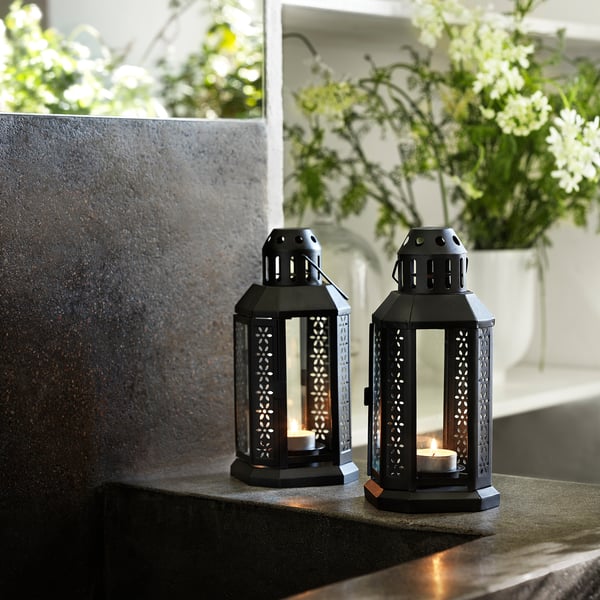 Two black GLIMMA lanterns with lit candles on a stone surface. One lantern has a tealight candle. Decorative, metallic design with cutouts.