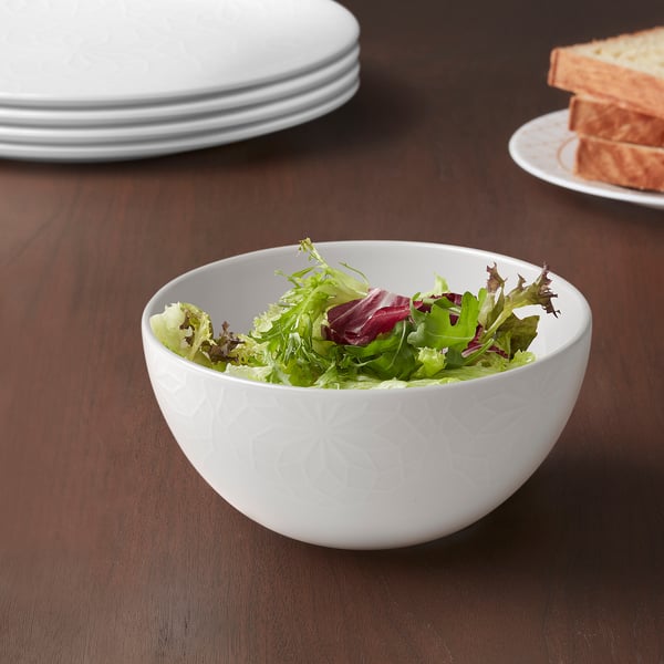 White bowl with green salad on wooden table, surrounded by plates and bread.
