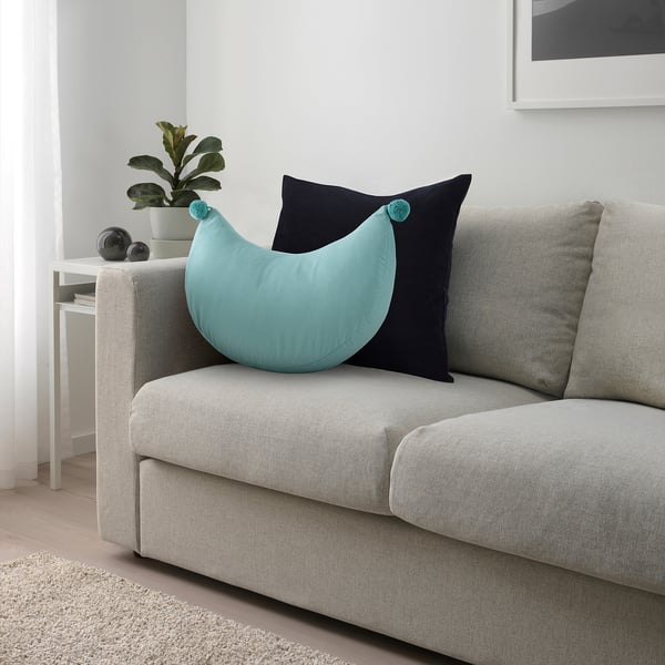 Turquoise crescent cushion with pompoms on light grey couch, beside small plant and larger black pillow.