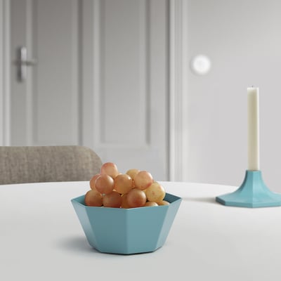 A small blue hexagonal bowl holds grapes. In a minimalist setting, the bowl sits on a white table near a turquoise candle holder.