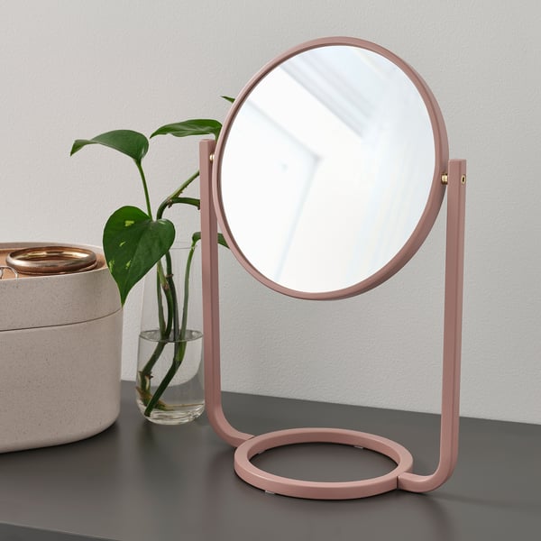 A pink table mirror with a magnifying side, displayed by a green plant on a shelf.