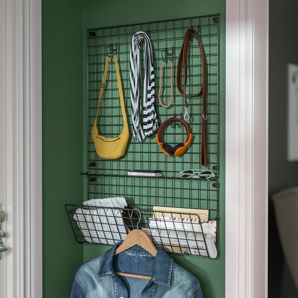 Wall organiser with mesh grids and baskets hangs on green wall. Holds accessories, bags, and denim jacket, showing efficient storage.