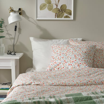 Floral pillow on bed beside lamp & plant, with botanical print. Cosy, light room.