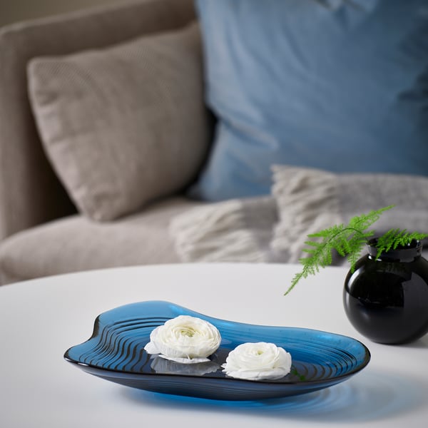 Blue glass bowl with flowers on white table, beside black vase with greenery.