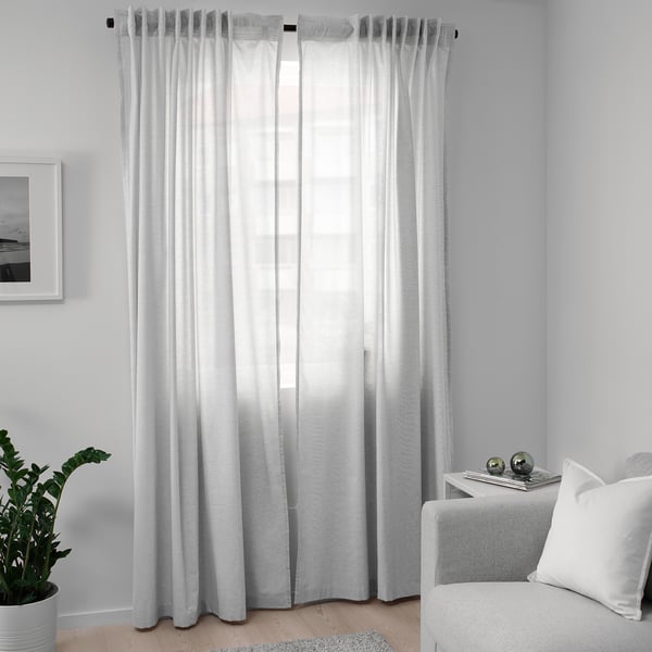 HANNALILL white curtains, made of 100% cotton, hung neatly beside a grey chair, creating a cosy atmosphere with soft lighting.