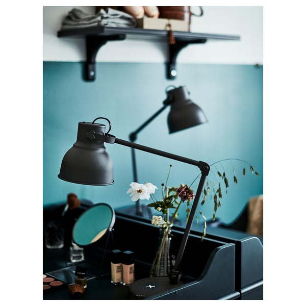 Modern bedroom with black adjustable HEKTAR lamps, one on a wall shelf and another on a desk beside a blue mirror and vase with flowers.