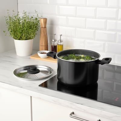 Black cookware with lid, boiling broccoli. Kitchen counter with herbs, seasonings, and digital stove.