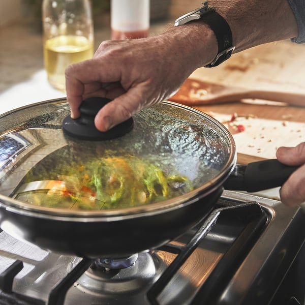 Person lifts lid from sizzling wok on stove, revealing colourful vegetables inside. The glass lid allows monitoring of cooking progress.