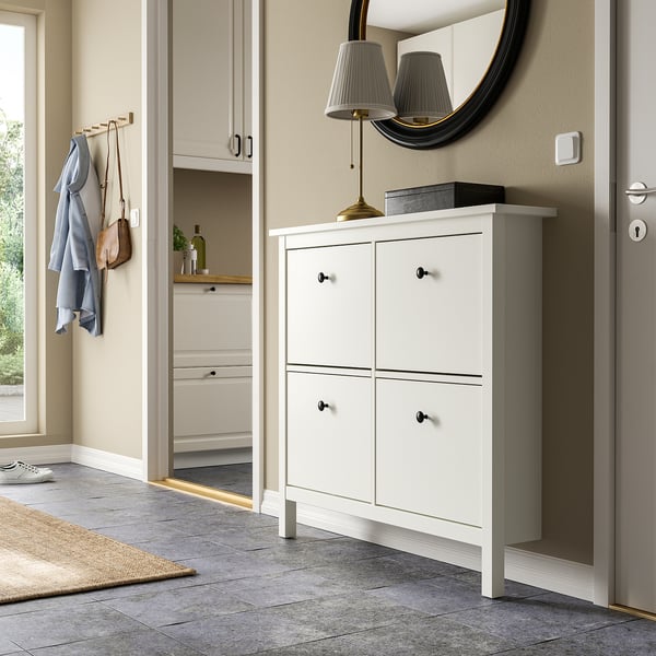 White HEMNES shoe cabinet, slim & tall, fits beneath wall-mounted mirror & next to coat hooks, with black handles & lamp on top.