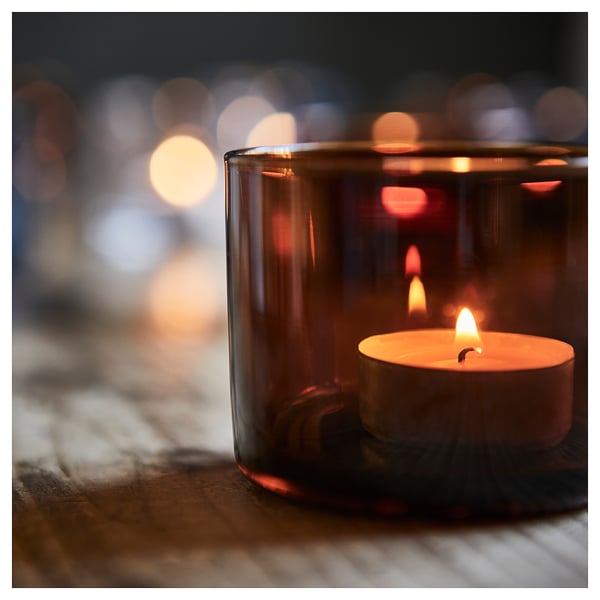 Small lit candle inside dark glass holder, creating warm glow, placed on textured surface.