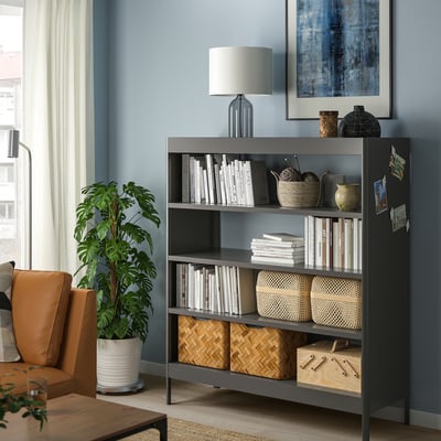 Black IDÅSEN bookshelf with adjustable shelves against blue wall, holding books and decor, near orange chair and plant.