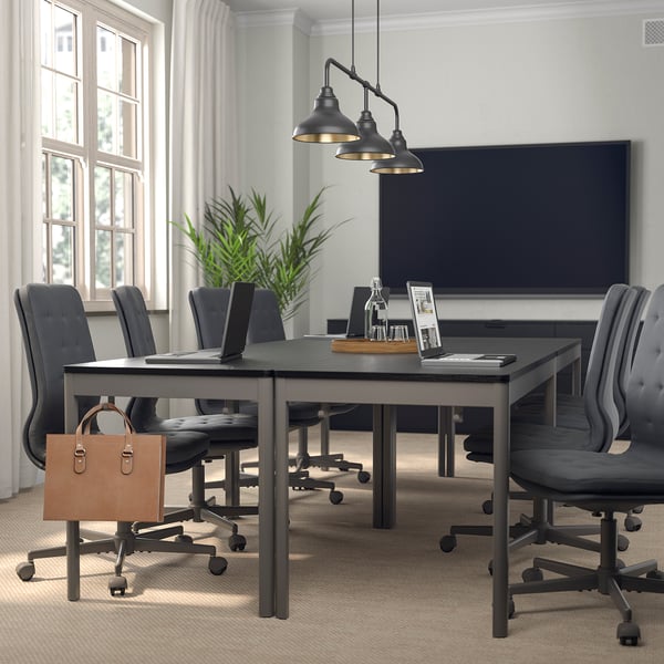 Modern meeting room with rectangular table, grey chairs, pendant lights, potted plant, and leather bag. Large windows.